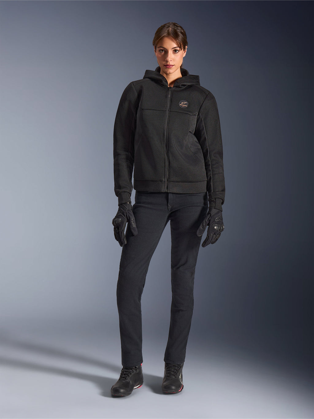 Kvindelig model iført Alpinestars Stella Chrome Superairflow Hoodie i sort, kombineret med sorte tekniske kørebukser og handsker, med 3D-mesh tekstilkonstruktion og kobberfarvet logo på brystet, urban sport-motorcykelbeklædning til kvinder.
