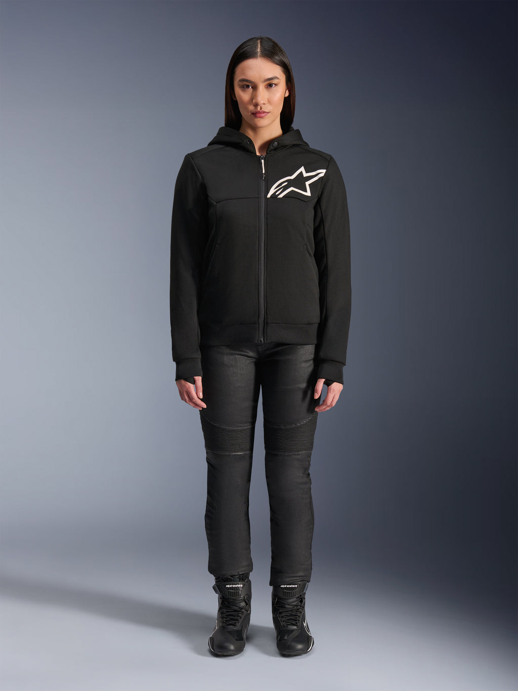 Kvinde iført Alpinestars Stella Chrome V2 Sport hættetrøje, sort med hvidt Astars-logo på brystet, kombineret med sorte tekniske motorcykelbukser og støvler, urbant motorcykeltøjsdesign