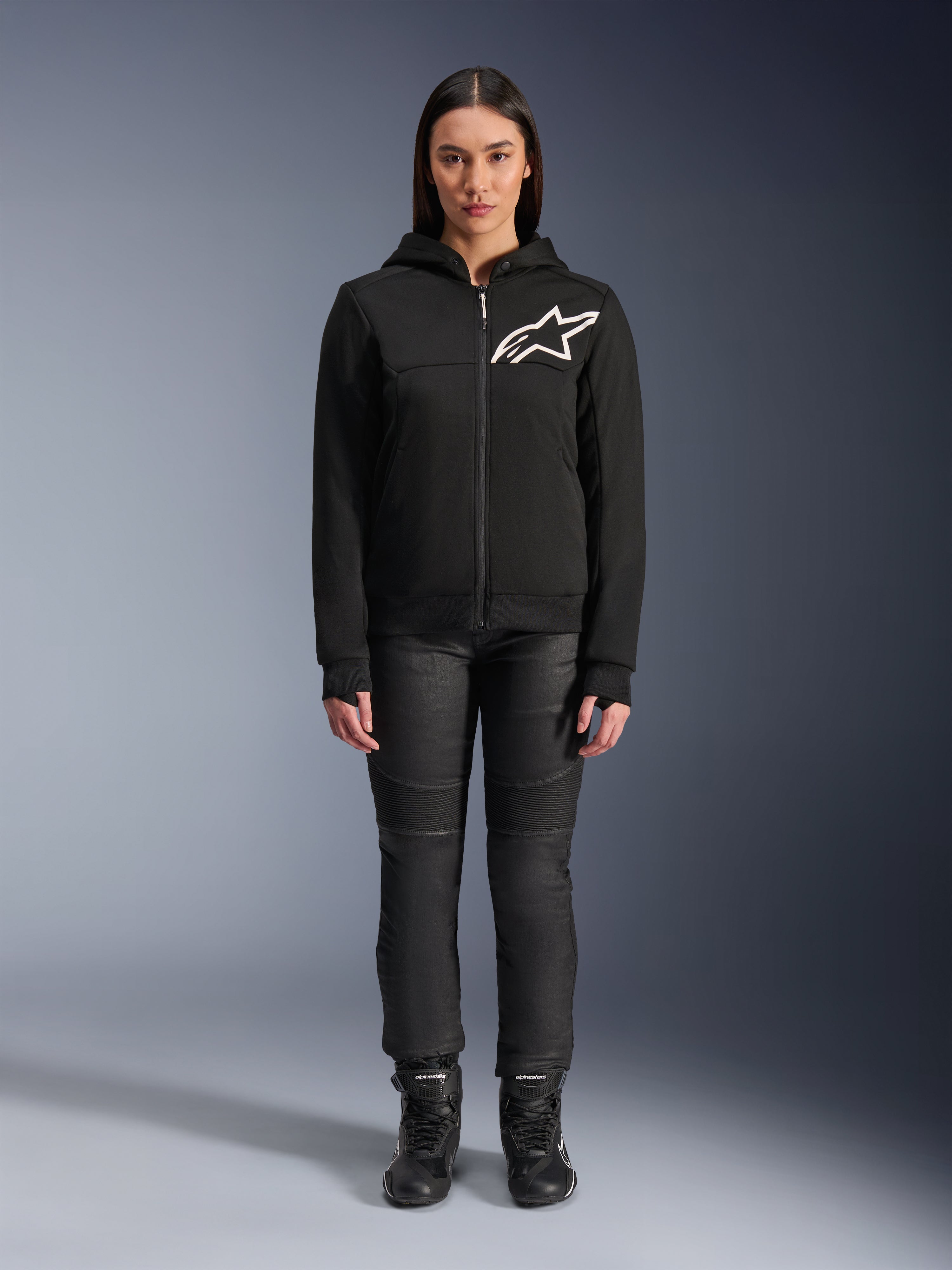Kvinde iført Alpinestars Stella Chrome V2 Sport hættetrøje, sort med hvidt Astars-logo på brystet, kombineret med sorte tekniske motorcykelbukser og støvler, urbant motorcykeltøjsdesign