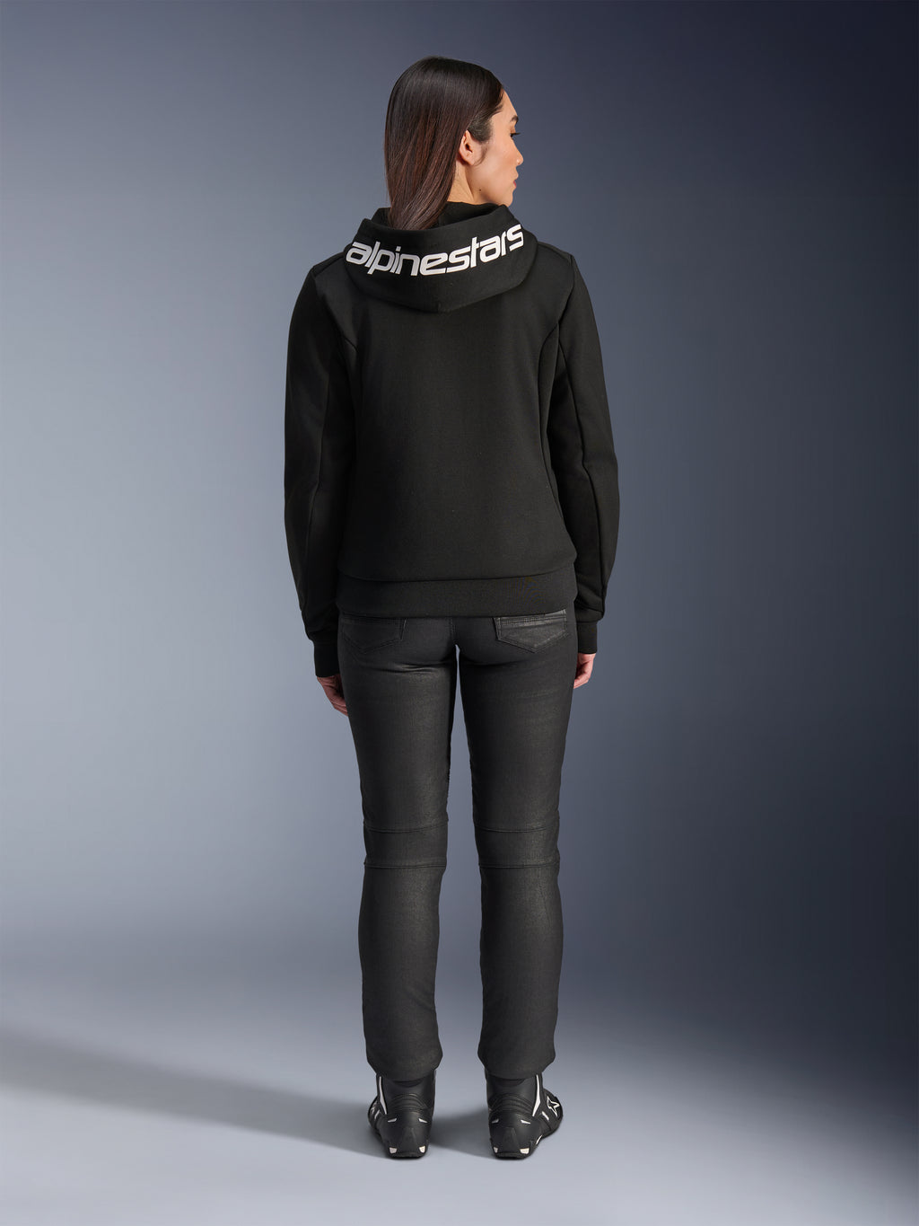 Set bagfra af en model iført Alpinestars Stella Chrome V2 Sport Hoodie i sort, med et markant hvidt Alpinestars-logo på hætten, kombineret med sorte motorcykelbukser og støvler, urban sport motorcykeludstyr til kvinder