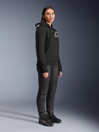 Kvinde iført Alpinestars Stella Chrome V2 Sport hættetrøje i sort, med hvidt Alpinestars-logo, kombineret med sorte motorcykelbukser og støvler, urbant køreudstyr til kvinder med en afslappet atletisk pasform
