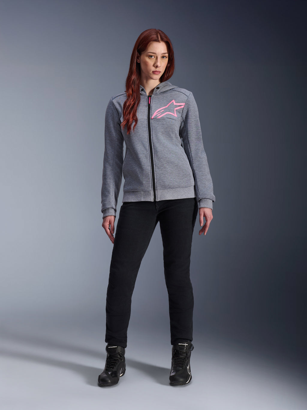 Alpinestars Stella Chrome V2 Sport Hoodie, Gray Heather Pink Fluo, båret af en kvinde med sorte bukser og støvler, med et lyserødt Alpinestars-stjernelogo på brystet, urbant motorcykeludstyr med gennemgående lynlås, letvægts tech-fleece med en afslappet atletisk pasform.