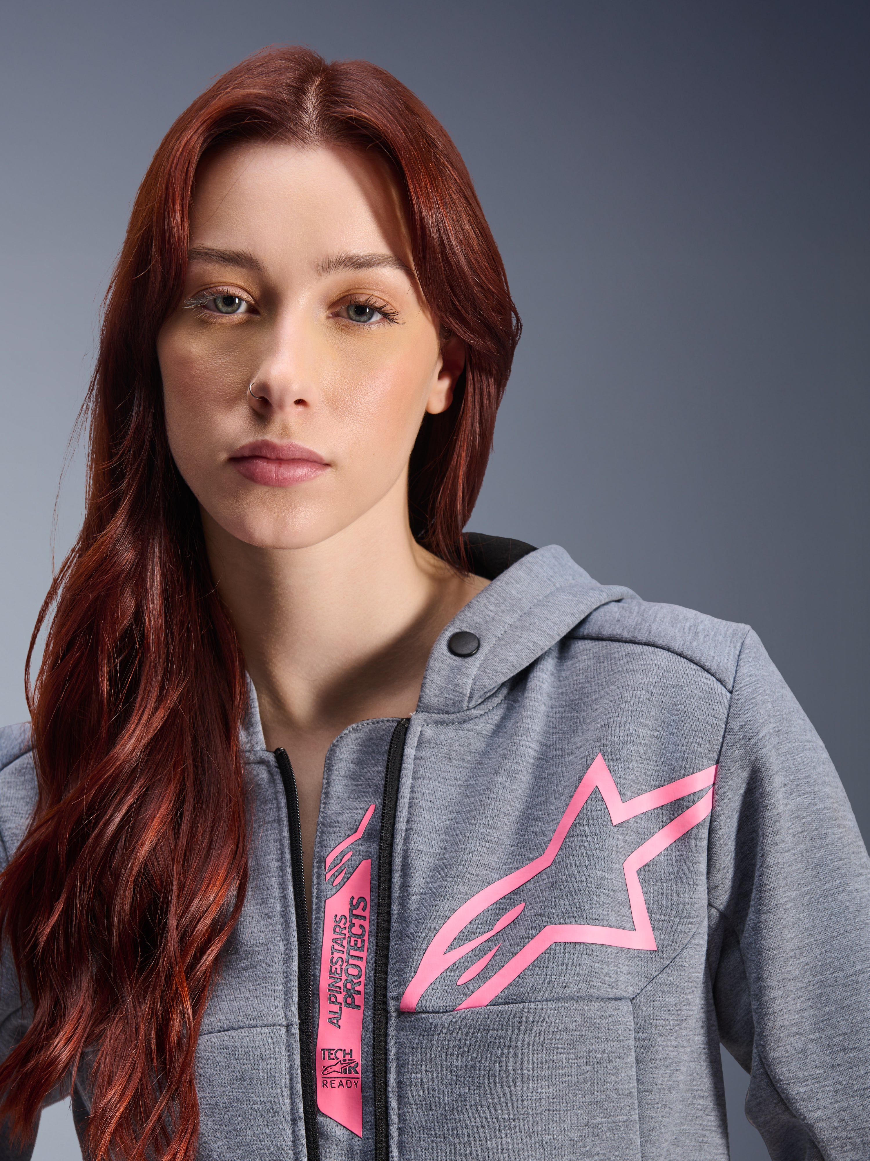 Kvinde iført Alpinestars Stella Chrome V2 Sport hættetrøje, Gray Heather Pink Fluo, nærbillede af det lyserøde Alpinestars stjernelogo og Tech-Air Ready-branding, urban motorcykelbeklædning