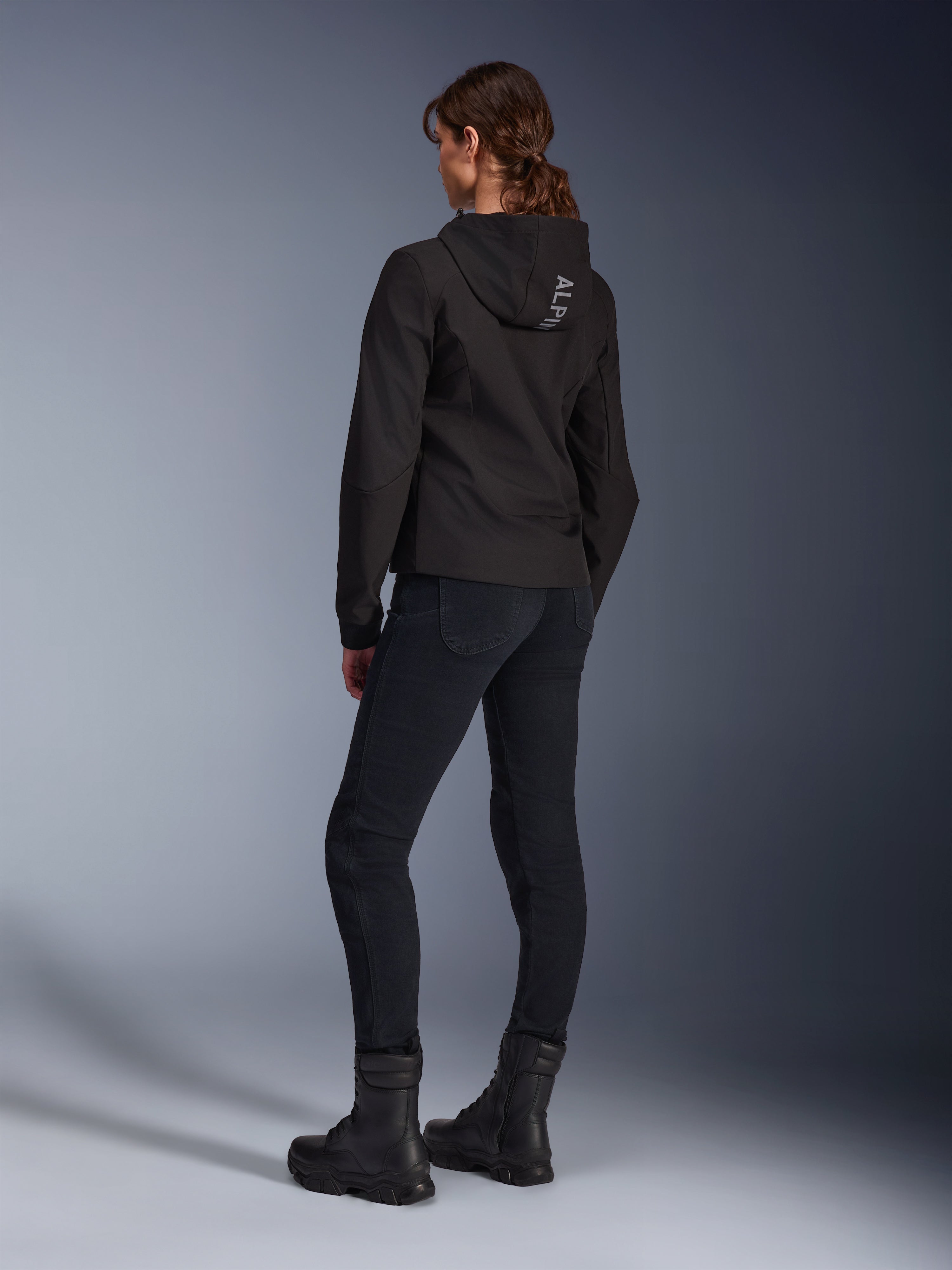 Alpinestars Stella Radium Tech Hoodie, sort urban kørehættetrøje til kvinder, set bagfra med 'ALPIN'-branding på hætten, kombineret med sorte tekniske kørebukser og sorte støvler, letvægts vindtæt tech-shell