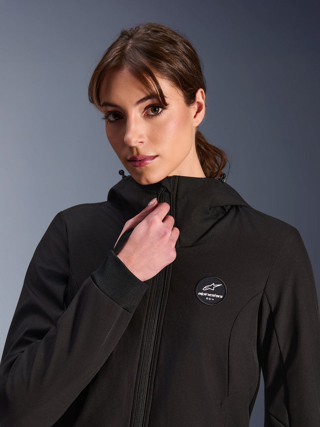 Woman Stella Radium Tech Hoodie, sort, båret af en kvinde, der justerer lynlåsen foran, cirkulært Alpinestars-logomærke på brystet, ribbede stretch-manchetter, vindtæt tekstilkonstruktion til bykørsel