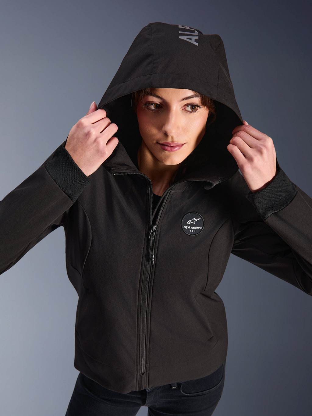 Kvinde iført Alpinestars Stella Radium Tech Hoodie i sort, urban motorcykel-hættetrøje med hætten oppe, cirkulært Alpinestars-logo på brystet, ribstrikkede manchetter og vindtæt 3-lags konstruktion.