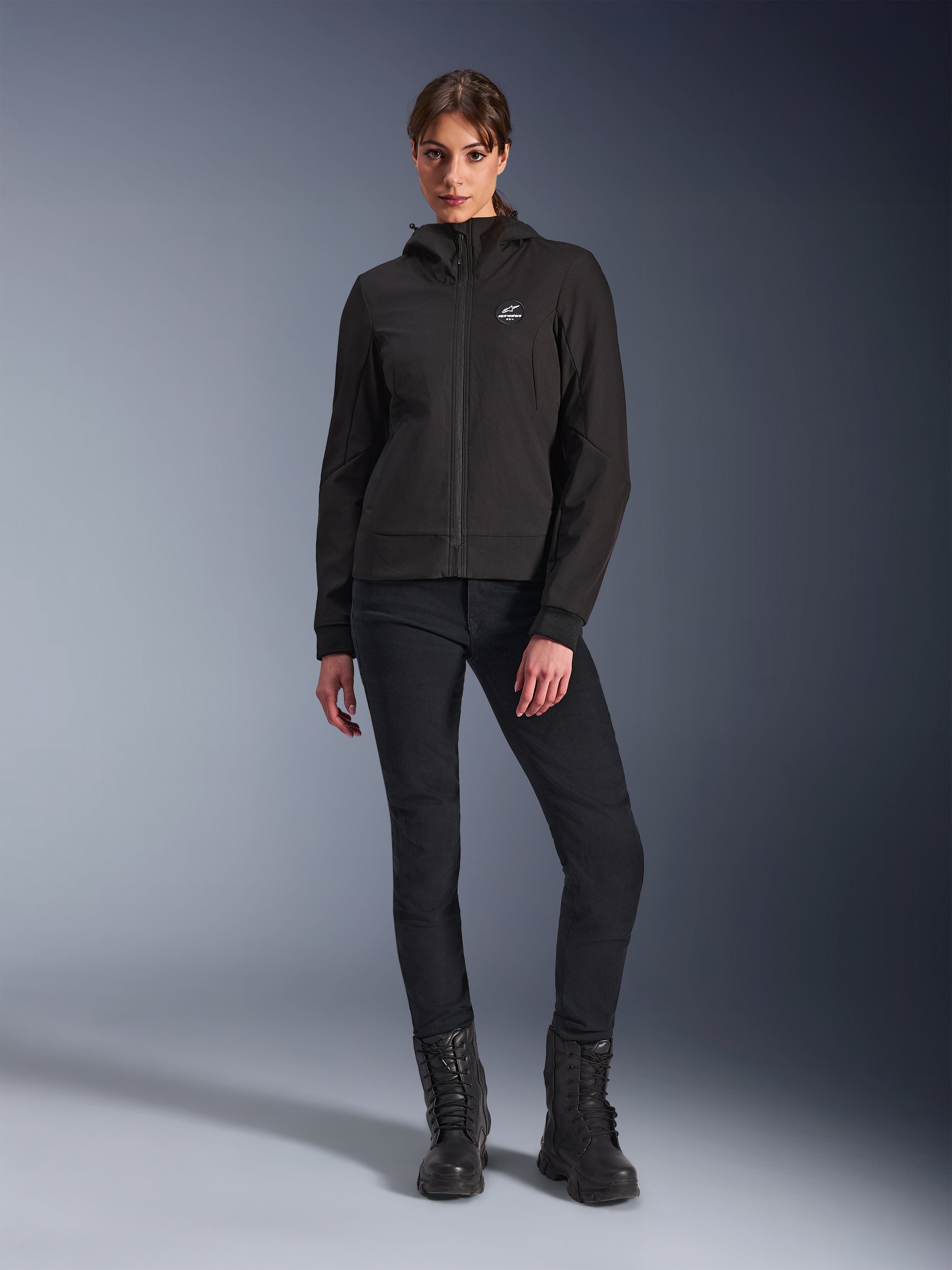 Alpinestars Stella Radium Tech Hoodie i sort, urban motorcykel-tech shell til kvinder, båret med sorte slim-fit bukser og sorte støvler, med et lille logo på brystet og hætte, letvægts vindtæt konstruktion til bykørsel