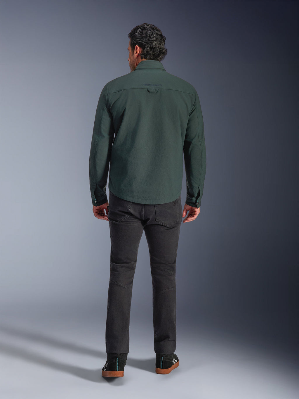 Alpinestars Bryon vandafvisende overshirt i Dark Emerald, set bagfra på model, kombineret med mørkegrå motorcykelbukser og sorte køresko, viser et klassisk arbejdstøjsinspireret design med en lidt længere ryg for bedre dækning under kørsel.