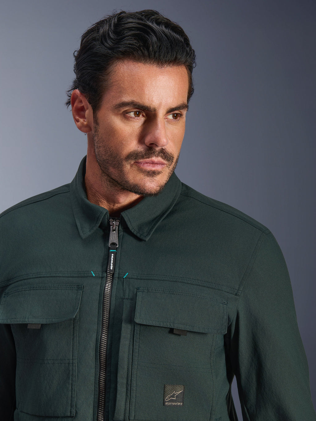 Alpinestars Bryon vandafvisende overshirt i Dark Emerald, nærbillede af en mand iført den urbane motorcykeljakke, tekstur af vokset lærred, frontlynlås med diskrete blågrønne accenter, brystlommer og logo-patch.