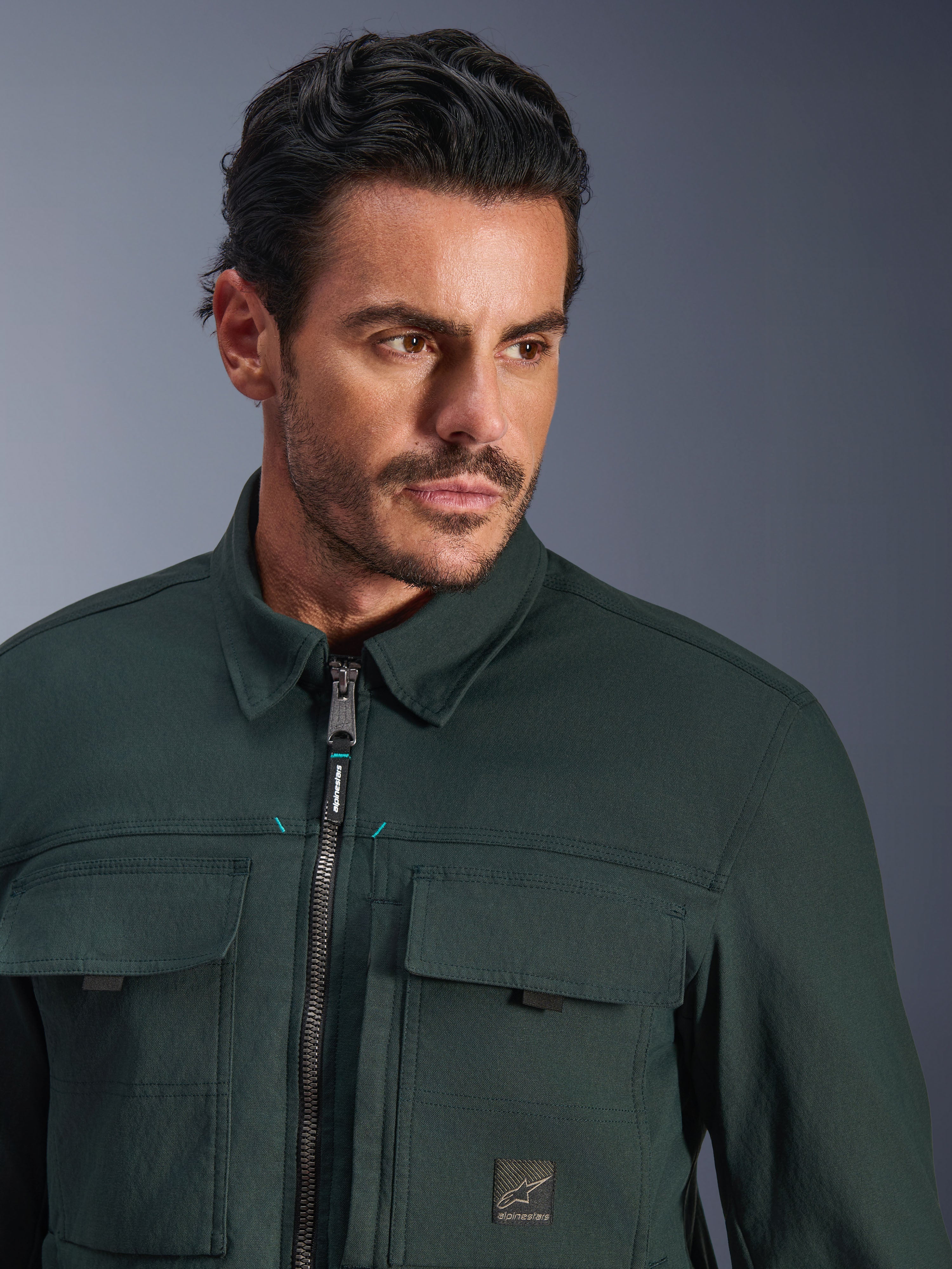 Alpinestars Bryon vandafvisende overshirt i Dark Emerald, nærbillede af en mand iført den urbane motorcykeljakke, tekstur af vokset lærred, frontlynlås med diskrete blågrønne accenter, brystlommer og logo-patch.