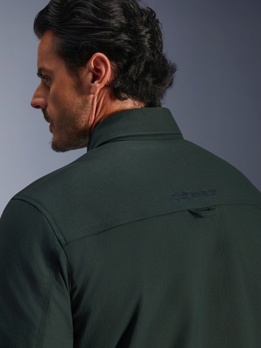 Alpinestars Bryon Water Resistant Overshirt i mørk smaragdgrøn, nærbillede af øverste del af ryg og krave på en mandlig model, med broderet Alpinestars-logo og lille stofstrop til ophæng, fremstillet i tekstureret vandafvisende vokset lærred til urban motorcykelkørsel.