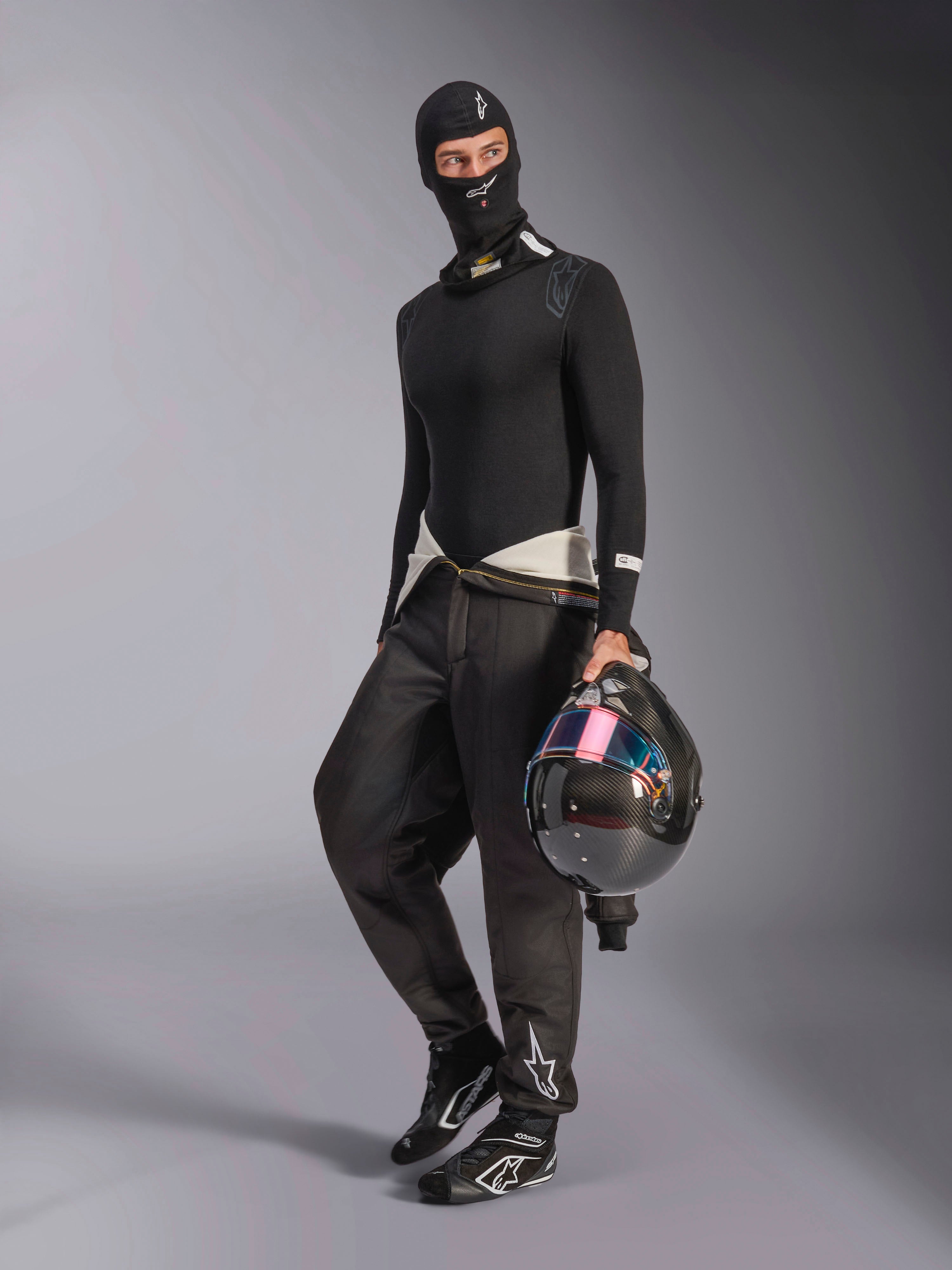 Alpinestars ZX Evo V3 Balaclava, Black, worn by a driver with complete auto racing base layer gear, featuring the white logo on the forehead, holding a carbon fiber helmet with an iridescent visor, professional fire-resistant motorsport apparel.