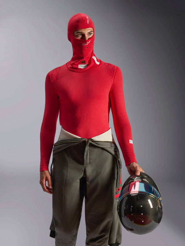 Alpinestars ZX Evo V3 Balaclava i rød, basislag til motorsport båret af en kører med matchende rød langærmet trøje og olivengrønne racerbukser, holder en kulfiberhjelm, professionelt flammehæmmende motorsportsudstyr
