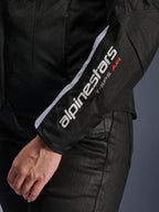 Alpinestars Stella T-SPS Air V2 jakke, motorcykeljakke til damer, sort og hvid, nærbillede af ærmet med hvidt Alpinestars-logo og T-SPS AIR-tekst, mesh- og tekstilkonstruktion for luftgennemstrømning, båret med sorte kørebukser