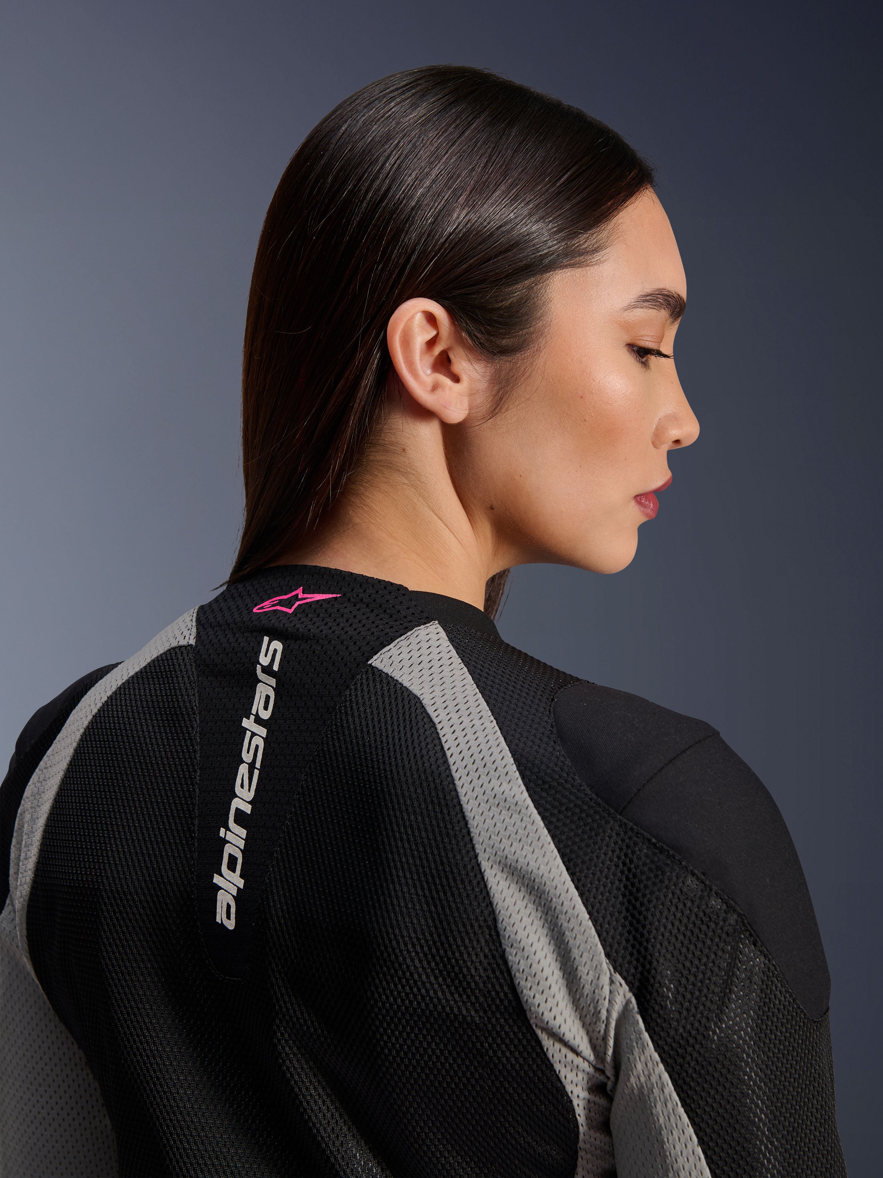 Nærbillede i profil af en kvinde iført Alpinestars Stella Troop-Air jakke, sort og askegrå mesh-konstruktion, lodret hvidt Alpinestars-logo på den øverste del af ryggen med et Pink Fluo stjernelogo, letvægts teknisk motorcykeludstyr til kvinder