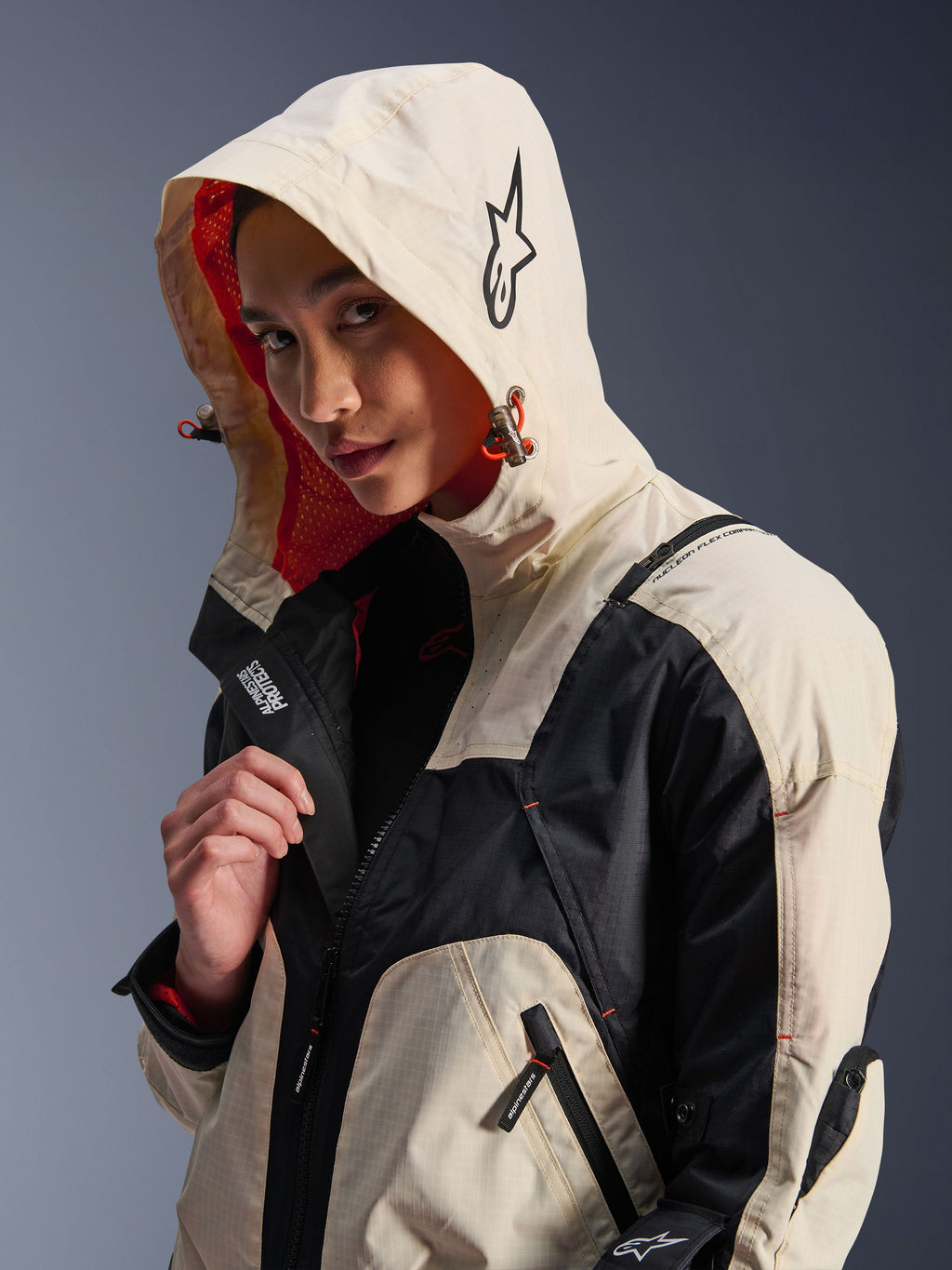 Alpinestars Molly Air-jakke i Black Almond, båret af en kvindelig model med hætten oppe, creme- og sort farveblokering, iøjnefaldende orange mesh-for, tekniske urbane designelementer, stjernelogo og lynlåsdetaljer