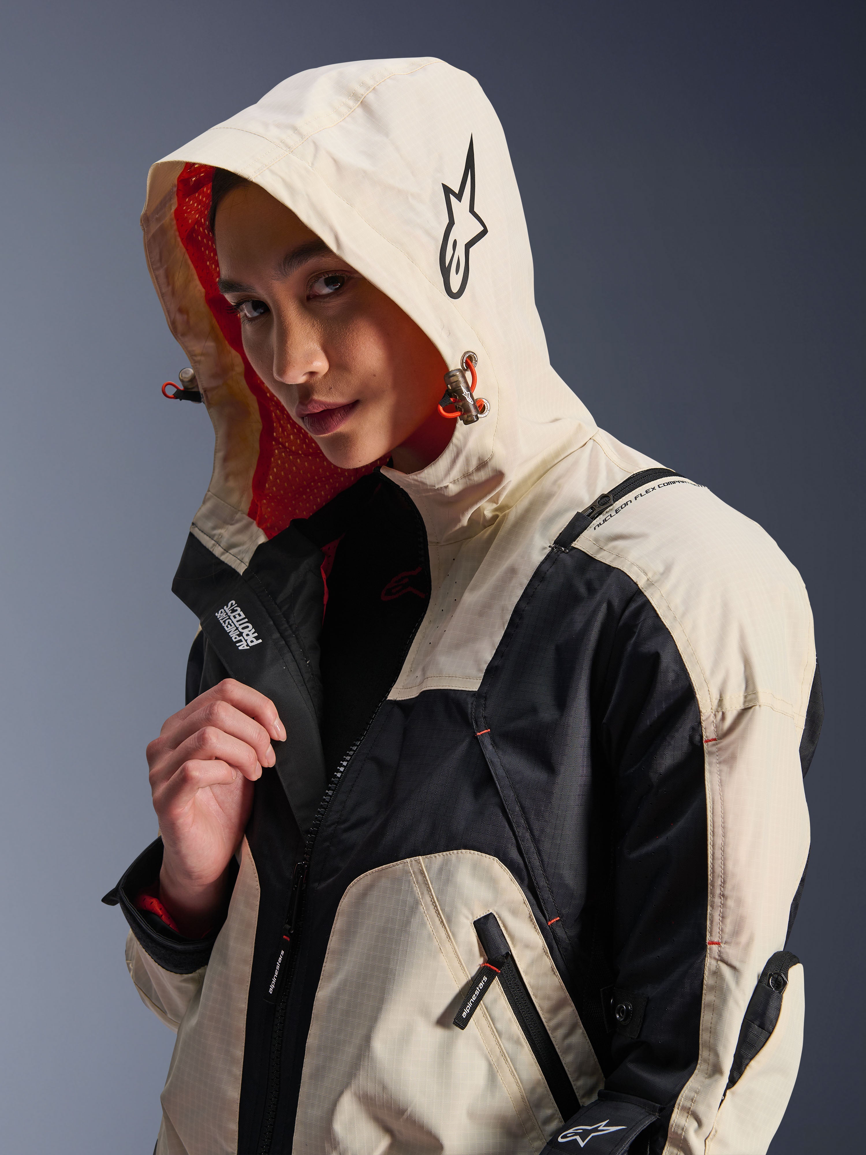 Alpinestars Molly Air-jakke i Black Almond, båret af en kvindelig model med hætten oppe, creme- og sort farveblokering, iøjnefaldende orange mesh-for, tekniske urbane designelementer, stjernelogo og lynlåsdetaljer