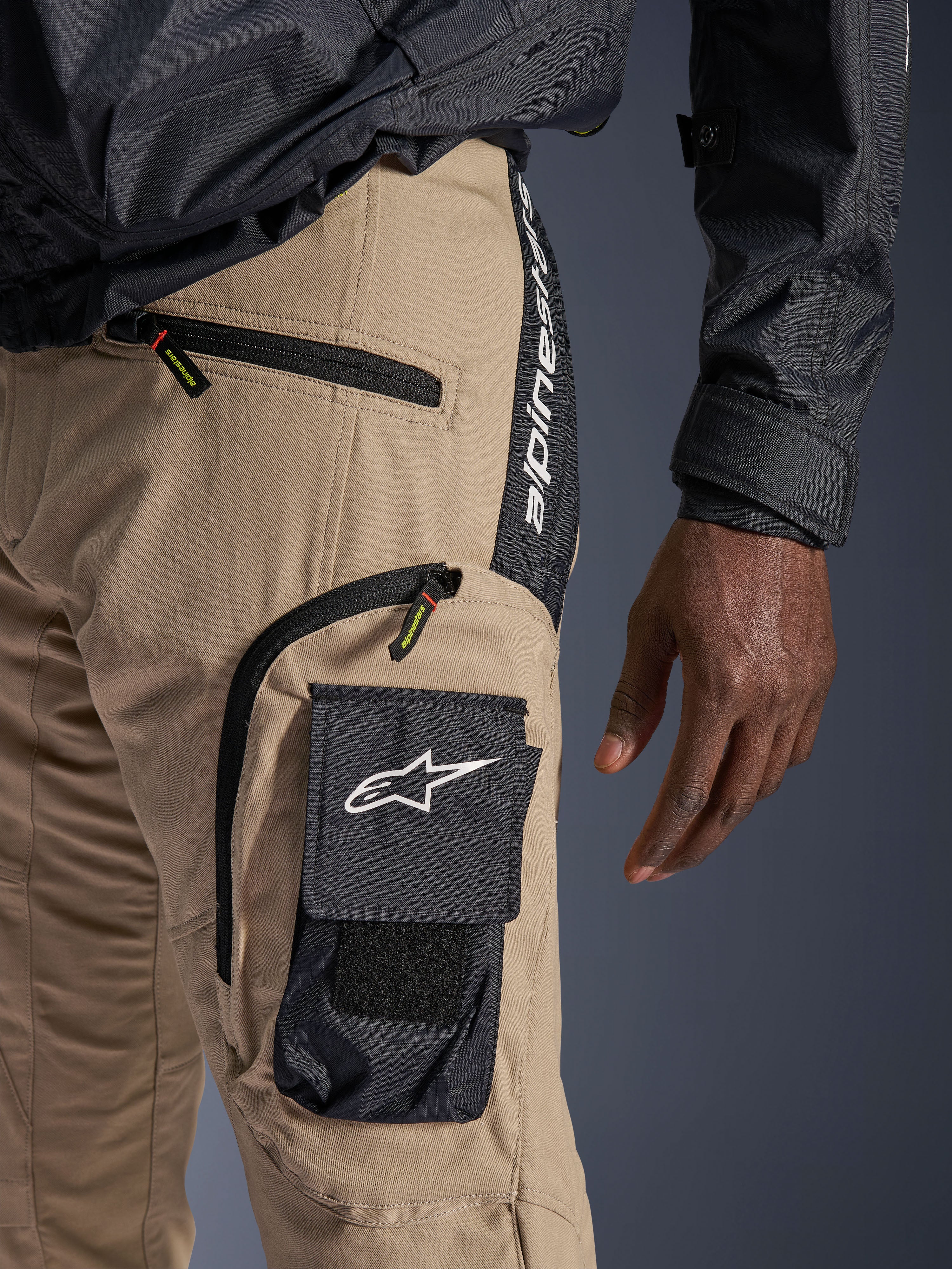 Alpinestars Acteon bukser i Sand, detaljeret visning af hofte- og lårområdet, med en sort cargolomme med hvidt stjernelogo, lynlåslommer og lodret Alpinestars-branding på et sort sidepanel, urban motorcykelbeklædning.