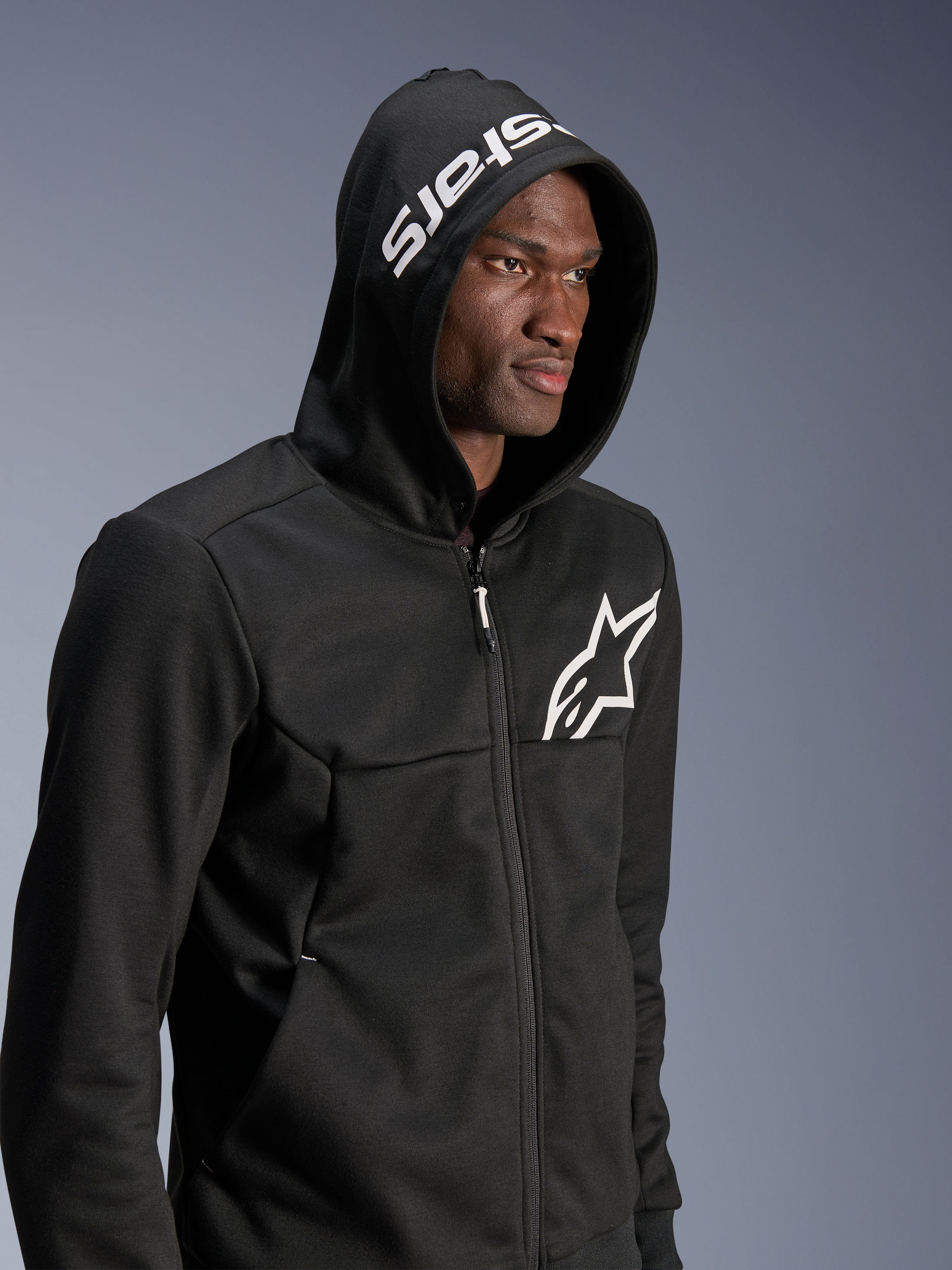 Alpinestars Chrome V2 Sport Hoodie, sort, urban motorcykel-hættetrøje båret af model med hætten oppe, hvidt stjerne-logo på brystet og hvidt stars-branding på hætten, tech fleece-konstruktion, afslappet motorcykel-streetwear