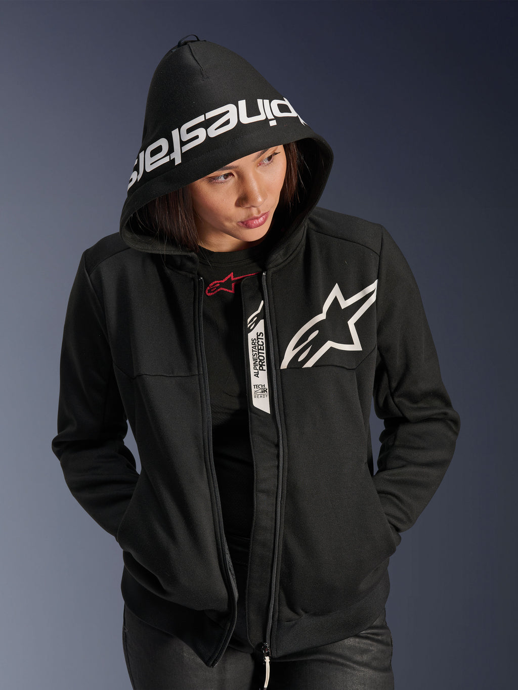 Alpinestars Stella Chrome V2 Sport hættetrøje til damer, sort og hvid, motorcykel-hættetrøje med hvidt stjernelogo på brystet og Alpinestars-branding på hættekanten, kvinde med hætten oppe, afslappet urban sportsstil med Tech-Air Ready-detaljer.