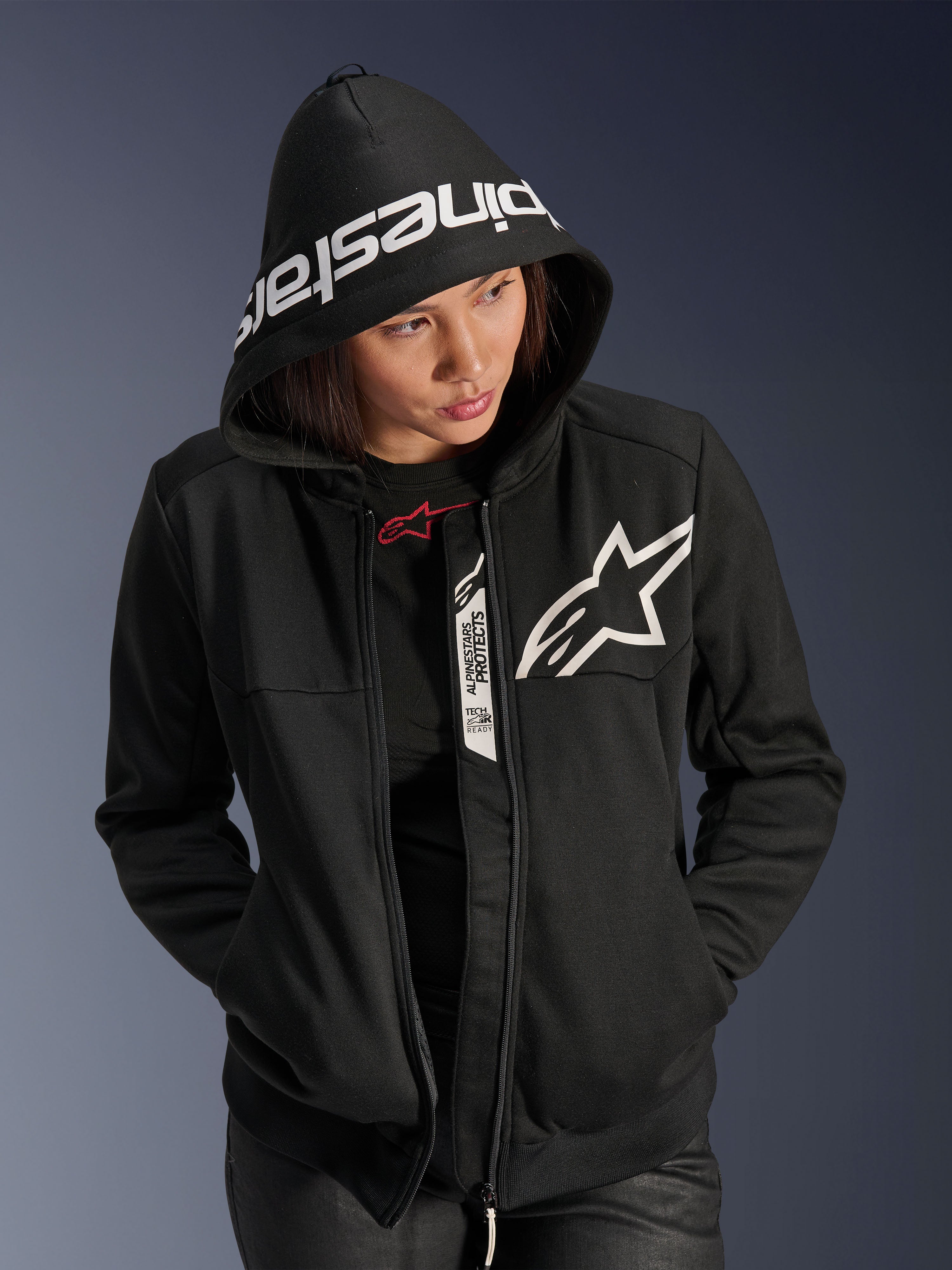 Alpinestars Stella Chrome V2 Sport hættetrøje til damer, sort og hvid, motorcykel-hættetrøje med hvidt stjernelogo på brystet og Alpinestars-branding på hættekanten, kvinde med hætten oppe, afslappet urban sportsstil med Tech-Air Ready-detaljer.