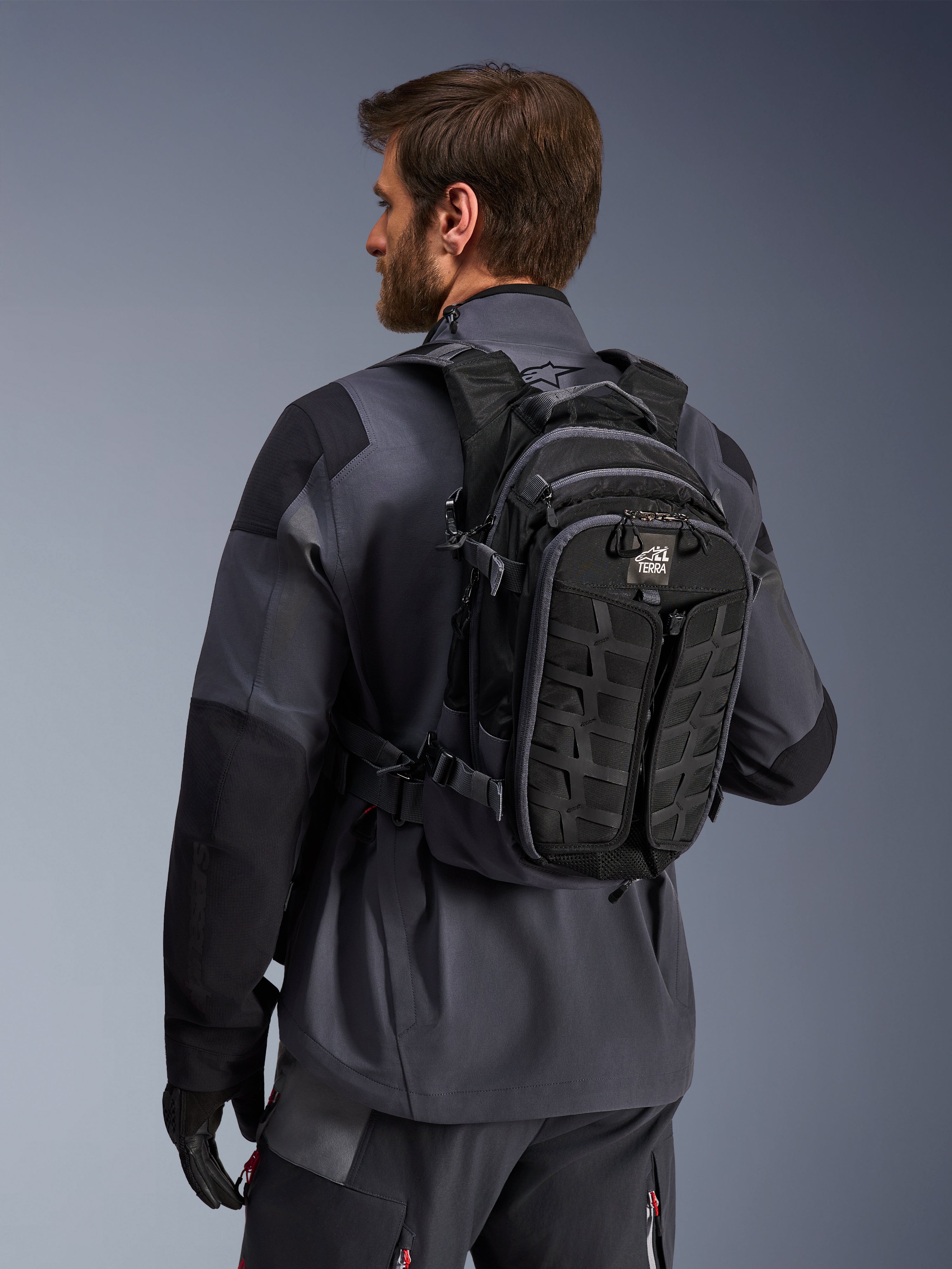 Alpinestars AT-15 rygsæk, adventure motorcykelrygsæk, sort og mørkegrå, båret af en kører i en grå jakke, med et teknisk design med MOLLE-webbing og 'TERRA'-branding, ergonomisk off-road udstyr