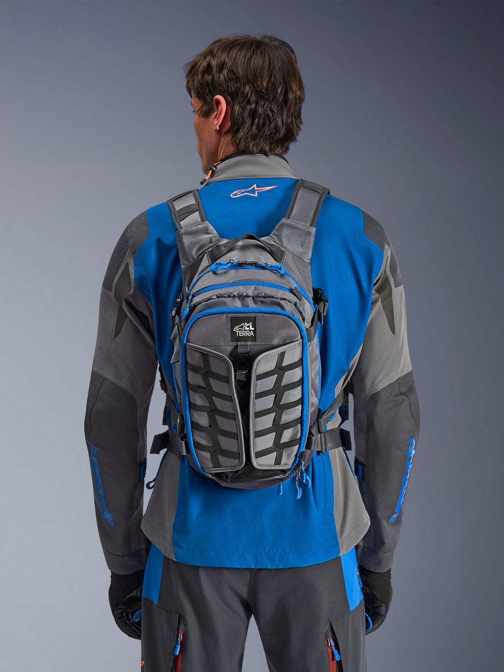 Alpinestars AT-15 Back Pack, adventure motorcykelrygsæk, mørkegrå og Deep Gray, bæres af en kører over en blå og grå jakke, med All Terra-branding, ergonomisk strukturelt design, justerbare stropper og blå lynlåsdetaljer.
