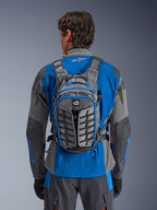 Alpinestars AT-15 Back Pack, adventure motorcykelrygsæk, mørkegrå og Deep Gray, bæres af en kører over en blå og grå jakke, med All Terra-branding, ergonomisk strukturelt design, justerbare stropper og blå lynlåsdetaljer.