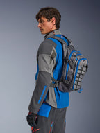 Alpinestars AT-15 Back Pack, adventure-rygsæk til motorcykel, mørkegrå med blå detaljer, bæres af en kører i koordineret gråt og blåt teknisk køreudstyr, med MOLLE-panel og robust konstruktion til off-road eventyr.