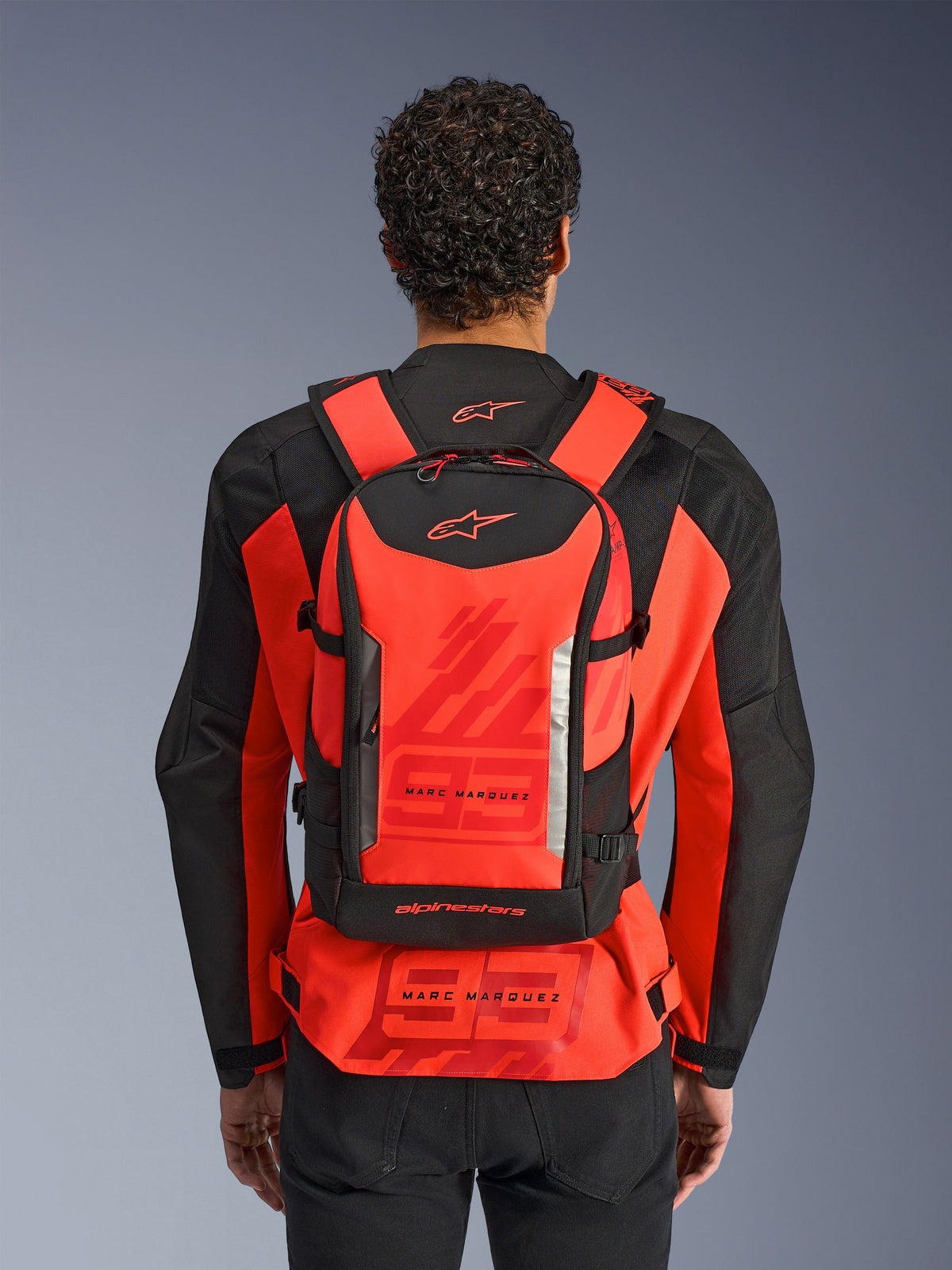 Alpinestars MM93 AMP3 rygsæk, teknisk motorcykelrygsæk, rød fluo og sort, båret af en rytter set bagfra over en matchende rød og sort MM93 racerjakke, med Marc Marquez-branding og Alpinestars-logoer.