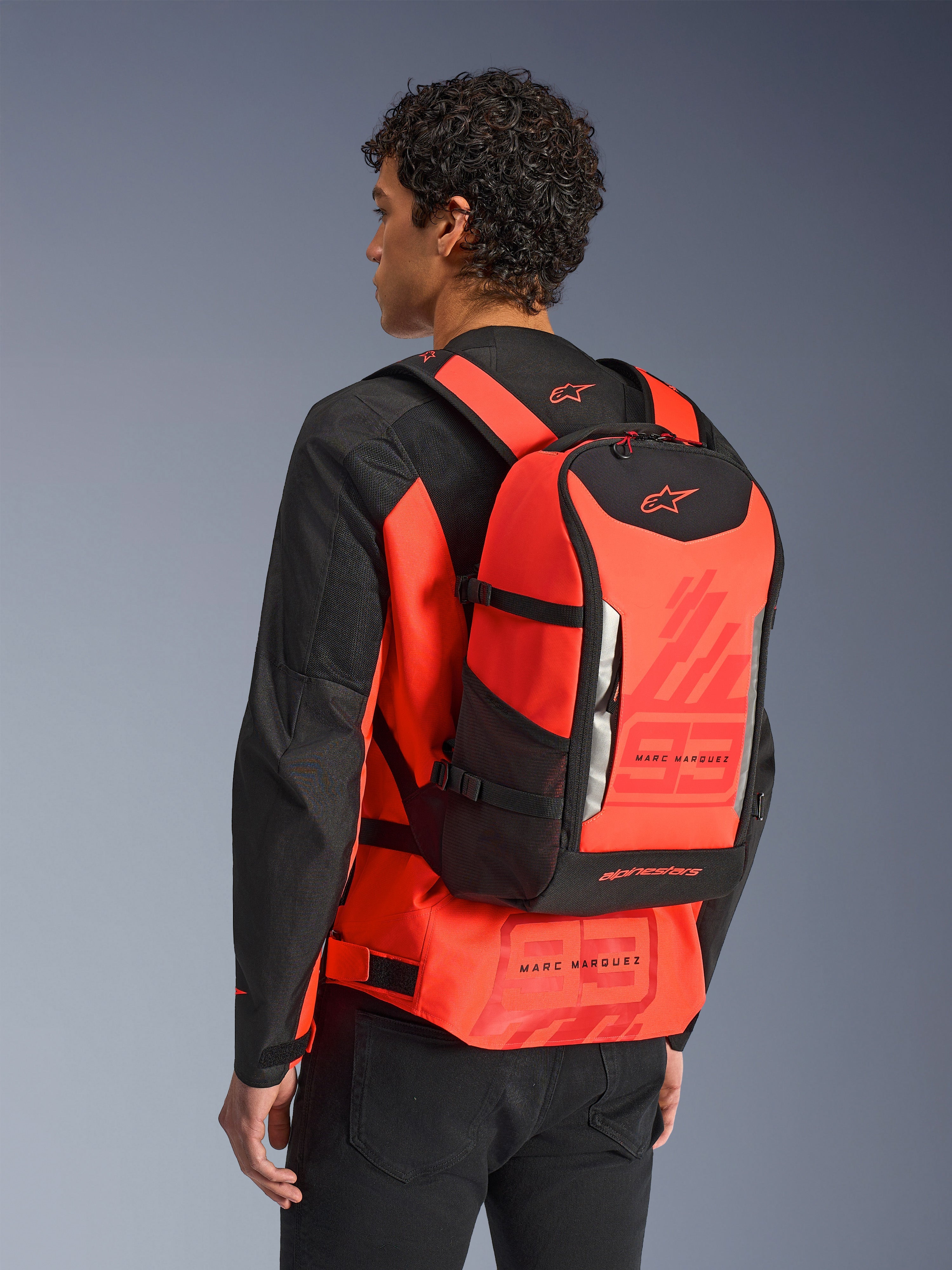 Alpinestars MM93 AMP3 rygsæk, teknisk motorcykelrygsæk, rød fluo og sort, bæres af en model bagfra over en matchende rød og sort jakke, med Marc Marquez '93'-grafik, justerbare stropper og strømlinet sportsinspireret design.