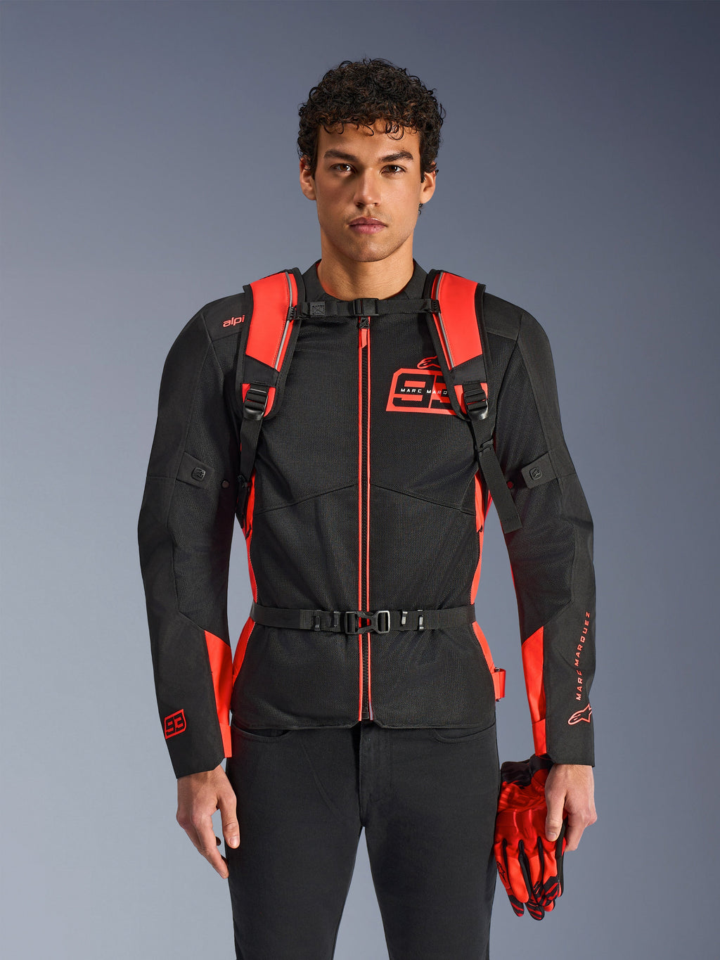 Mand iført Alpinestars MM93 AMP3 rygsæk, rød fluo og sort, med justerbare skulderstropper og brystrem med spænde, kombineret med en matchende MM93 motorcykeljakke og handsker, teknisk udstyr fra Marc Marquez-kollektionen