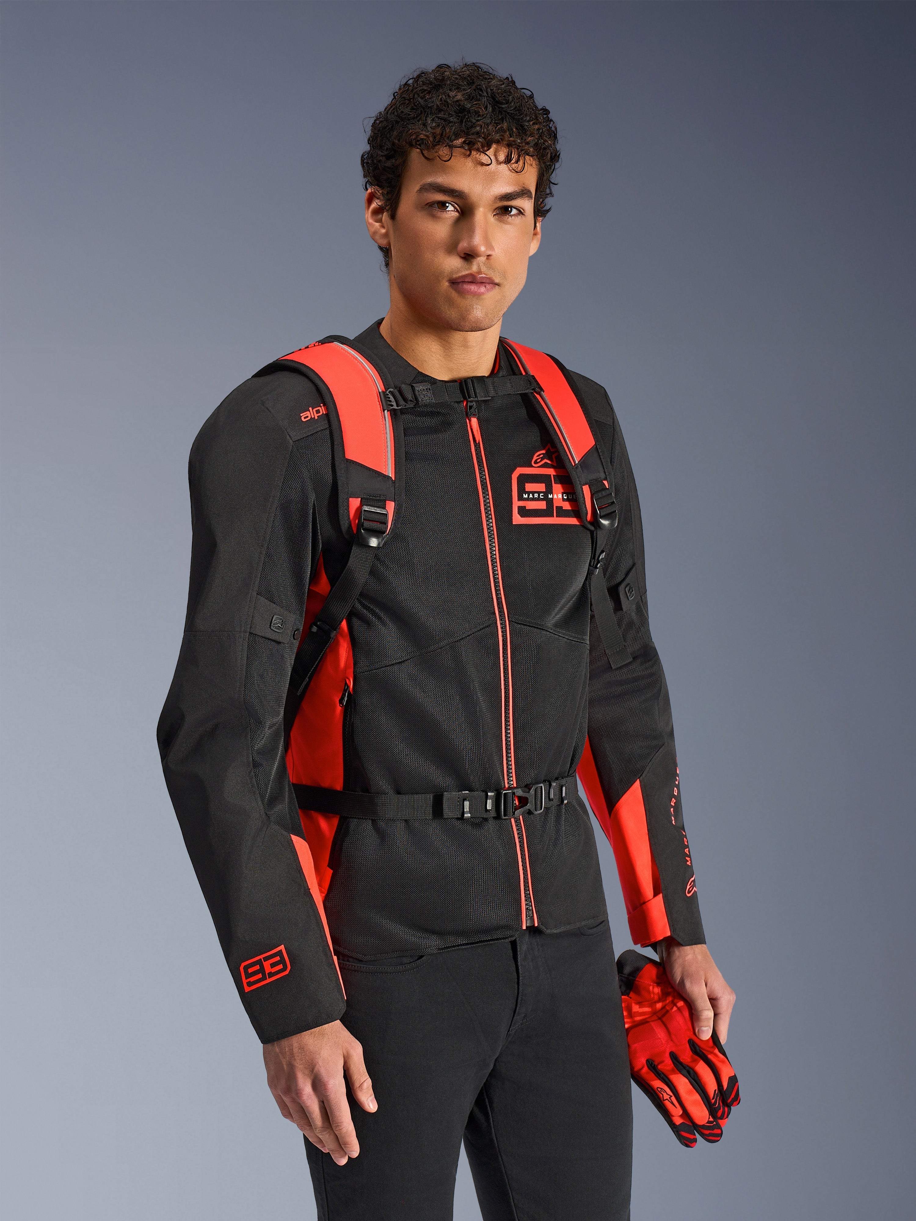 Alpinestars MM93 AMP3 rygsæk, rød fluo og sort, bæres af en model med en matchende sort og rød MM93 motorcykeljakke og handsker, set forfra med fokus på de justerbare skulderstropper og brystspænde, teknisk racing-inspireret udstyr