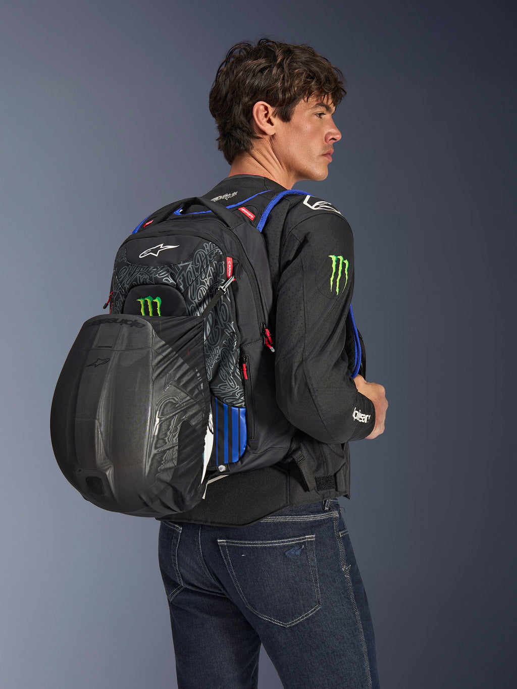 Alpinestars FQ20 City Hunter V2 Monster Rygsæk, urban motorcykelrygsæk i sort med rød fluo, blå og grønne detaljer, båret af en model med en sort hjelm fastgjort i det bageste net, stylet med en sort motorcykeljakke og blå jeans.