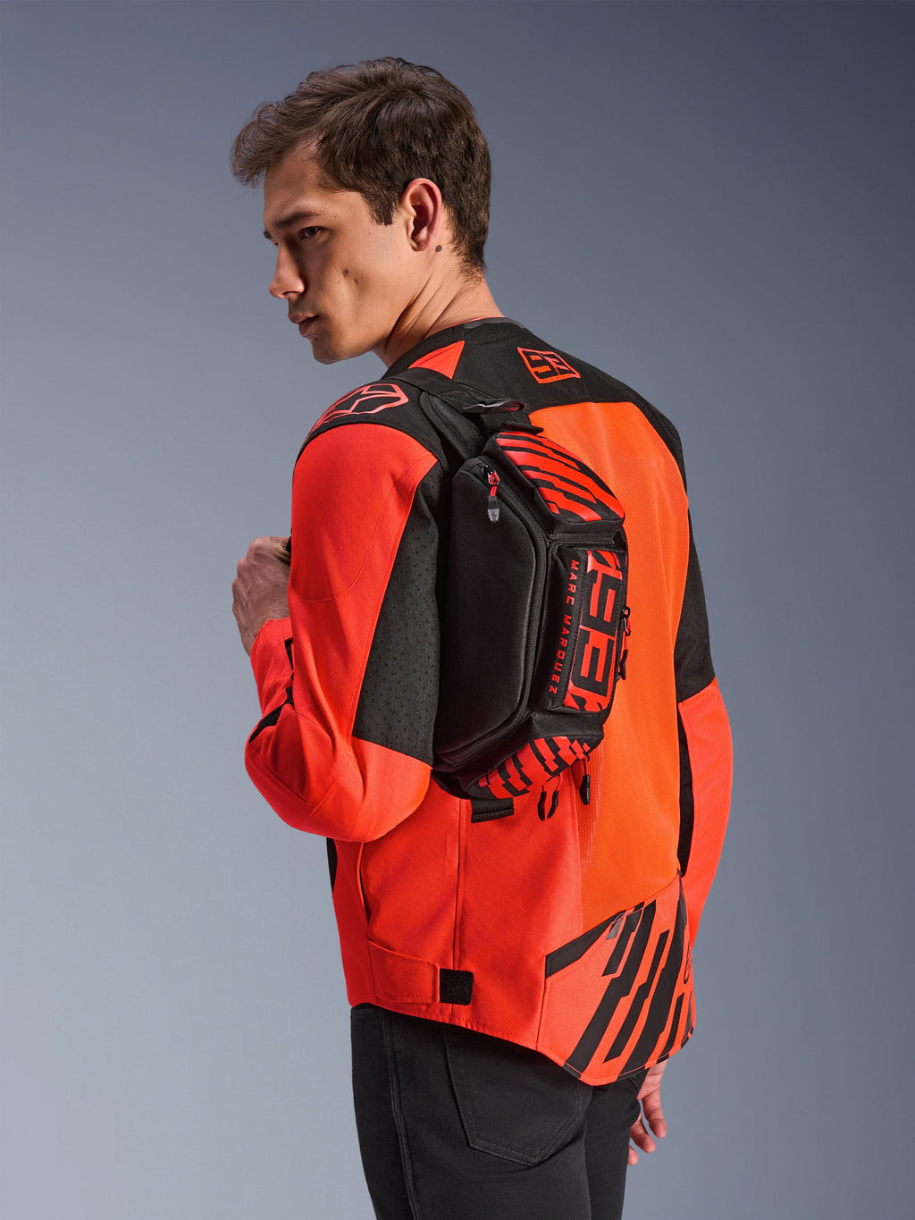 Alpinestars MM93 Waist Bag, sort med rød fluo og klar rød, båret af en model hen over ryggen over en rød og sort motorcykeljakke, med markant MM93-branding og Marc Marquez-grafik