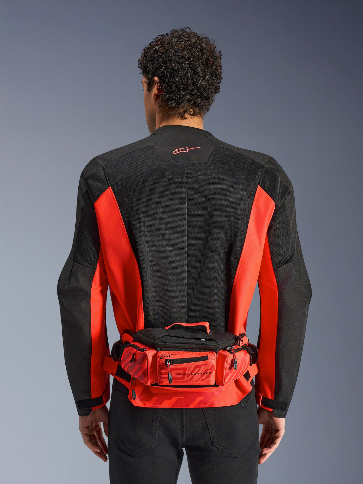 Alpinestars MM93 bæltetaske, rød og sort, båret over en sort og rød motorcykeljakke, med Marc Marquez 93-branding, justerbart bælte og rum med lynlås, funktionelt racer-inspireret tilbehør