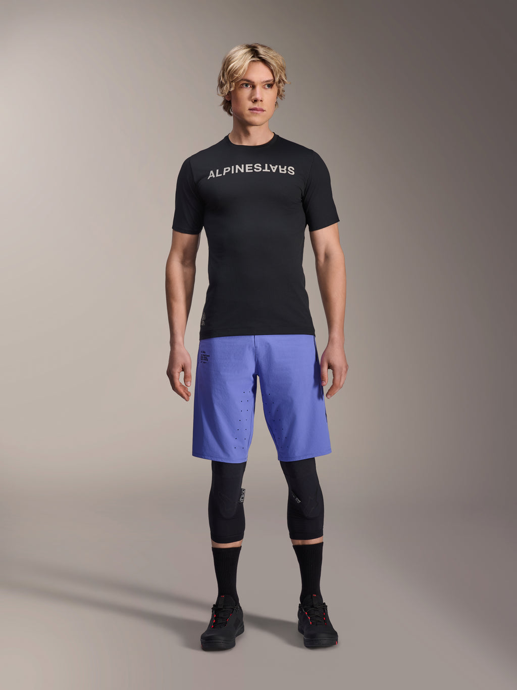 Alpinestars A-Aria Switch jersey, kortærmet MTB-trøje, sort med spejlvendt hvidt Alpinestars-logo, båret af en mandlig model med blå cykelshorts og sorte knæbeskyttere, åndbart performance-tøj til trail-kørsel
