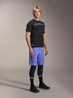 Mandlig model iført Alpinestars mountainbike-beklædning, herunder den sorte A-Aria Switch SS Jersey og Baja Blue A-Aria Lite shorts, kombineret med sorte knæbeskyttere, sorte sokker og sorte mountainbike-sko i et studiebillede i fuld figur.