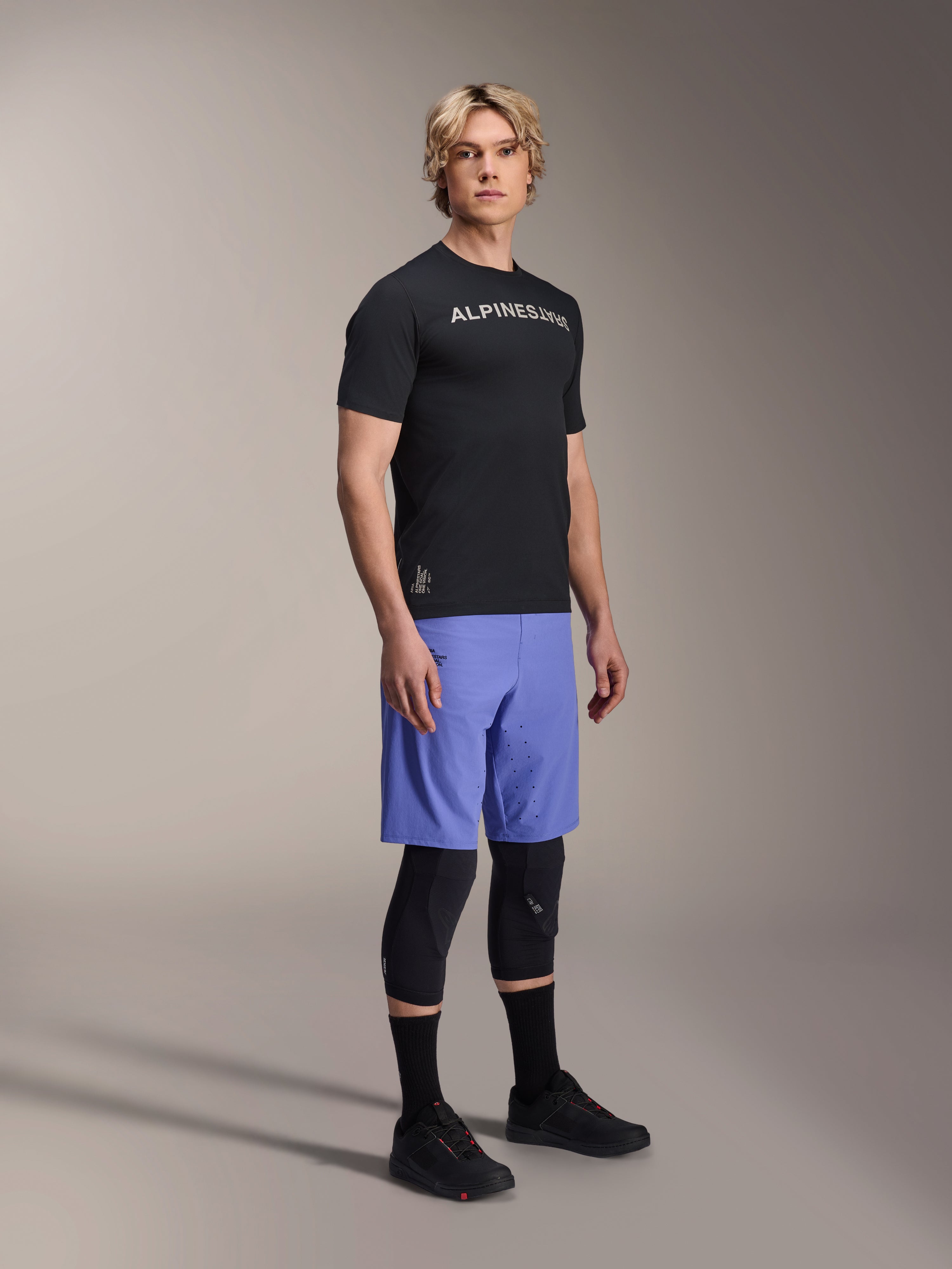 Mandlig model iført Alpinestars mountainbike-beklædning, herunder den sorte A-Aria Switch SS Jersey og Baja Blue A-Aria Lite shorts, kombineret med sorte knæbeskyttere, sorte sokker og sorte mountainbike-sko i et studiebillede i fuld figur.