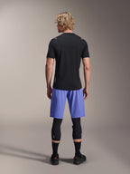 Mandlig rytter iført Alpinestars A-Aria Lite Shorts, MTB-shorts, Baja Blue, set bagfra, stylet med en sort kortærmet jersey, sorte knæbeskyttere og sorte cykelsko, performance mountainbike-beklædning