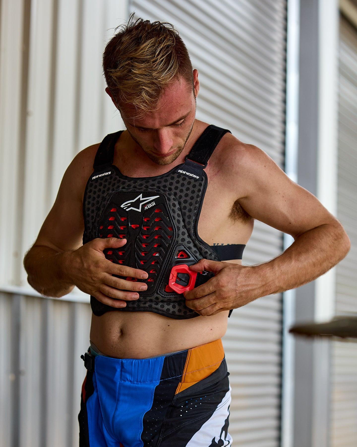Mand der tager Alpinestars A-5 Plasma brystbeskytter på, sort med røde detaljer og hvidt logo, bikageformet mesh-struktur, båret med blå og orange motocross-bukser, forberedelse til off-road kørsel