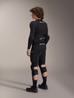 Model iført Alpinestars A-Impact Plasma Pro knæ- og skinnebensbeskyttere set bagfra, sort og hvid, der viser det tredobbelte strop-lukkesystem og ventilerede mesh-paneler, båret med et komplet sæt sort mountainbike-beskyttelsesudstyr, herunder et langærmet basislag og polstrede shorts