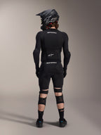 Alpinestars A-Impact Plasma Pro knæ- og skinnebensbeskyttere, sort, set bagfra på en rytter iført komplet mountainbike-beskyttelsesudstyr inklusive en full-face hjelm og kropsbeskyttelse, med tredobbelt strop-lukkesystem og ventilerede mesh-paneler for sikker pasform og luftgennemstrømning