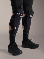 Alpinestars A-Impact Plasma Pro knæ- og skinnebensbeskyttere, sort og hvid, med hvidt Alpinestars-logo og tredobbelt strop-lukkesystem, vist med sorte sokker og sorte sportssko, slidstærk beskyttelse til off-road cykling
