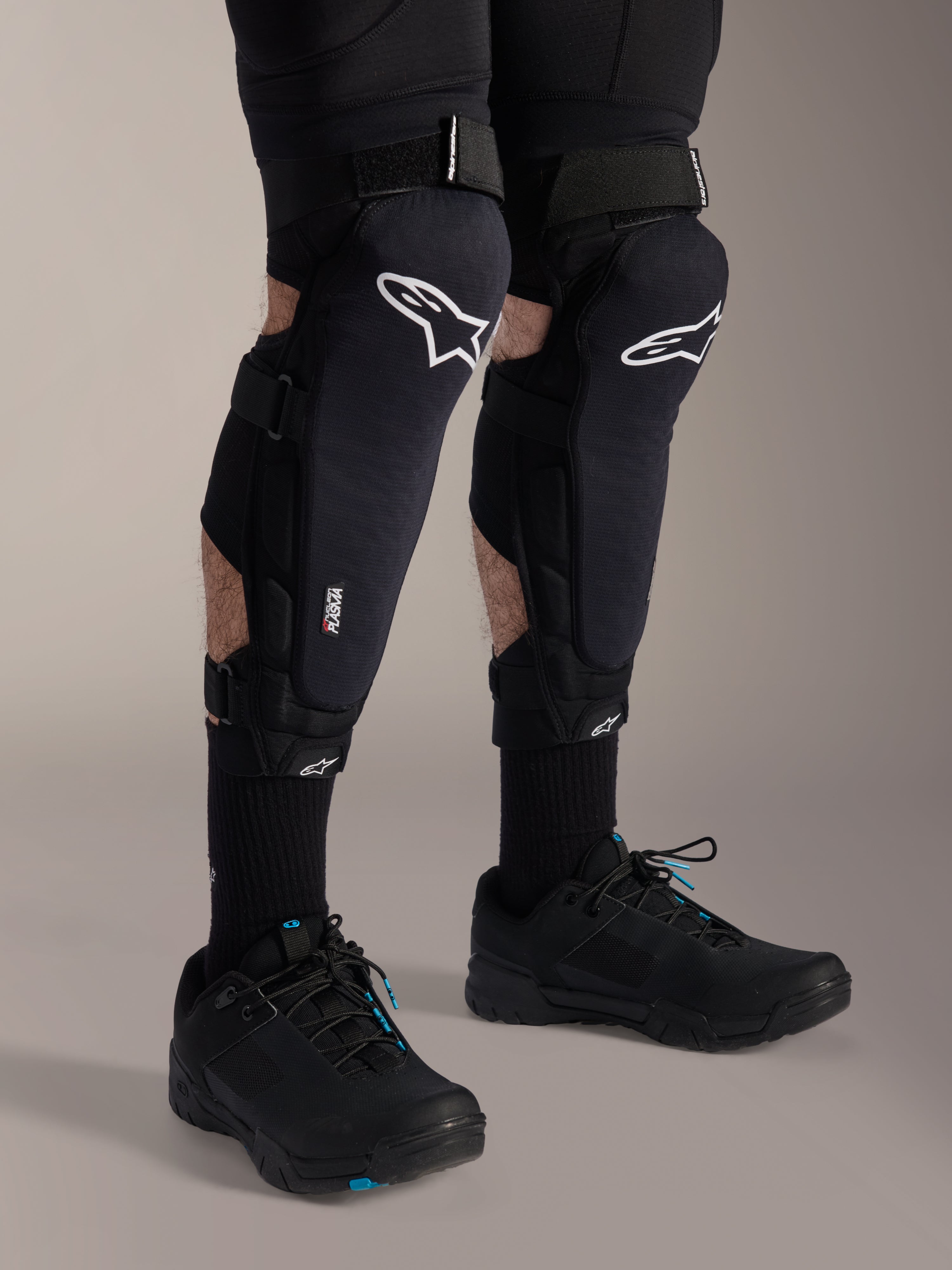 Alpinestars A-Impact Plasma Pro knæbeskyttere, sort og hvid, med Nucleon PLASMA-teknologi branding, båret med sorte strømper og sorte mountainbike-sko, sikre velcrostropper, ergonomisk beskyttelsesudstyr til off-road cykling