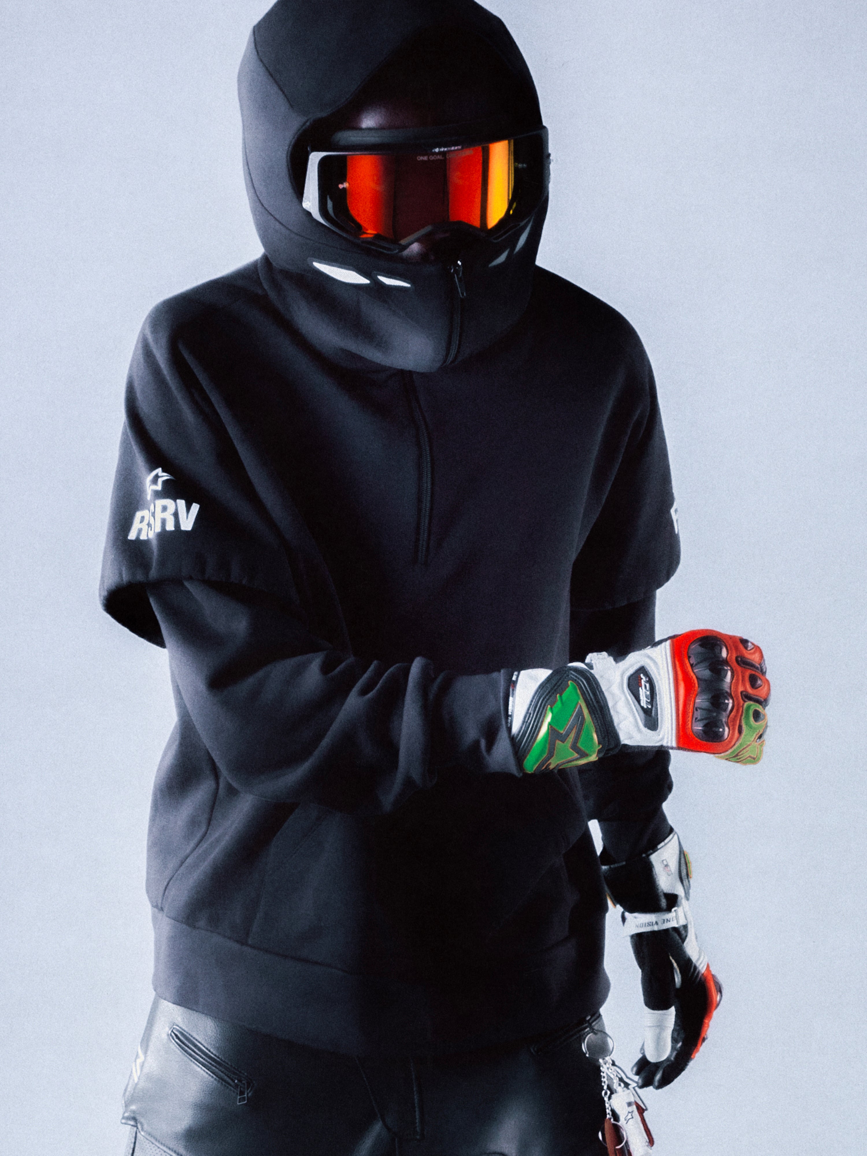 RSRV Vault Hoodie, Black Premium Streetwear Hoodie, Person wearing motorsport-inspired luxury hoodie with hood up, black helmet with reflective red-orange goggles, colorful racing gloves, sleek urban performance style