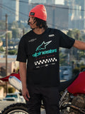 Alpinestars Aplomb Oversized CSF Tee, kortærmet T-shirt, sort, med et stort Astars-logo i blågrøn og hvid farveovergang og ternet flag-grafik, båret med en koralfarvet hue, stående ved siden af en rød motorcykel, afslappet lifestyle streetwear