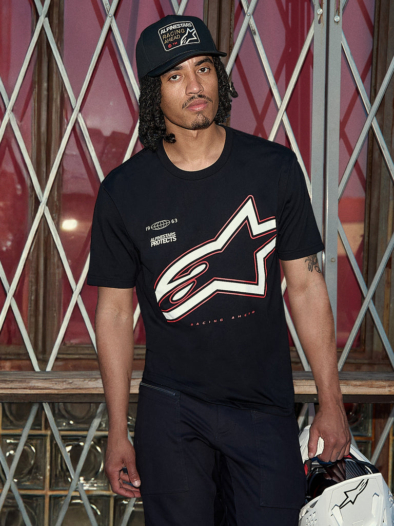 Alpinestars Authority CSF Tee, kortærmet t-shirt, sort, stort hvidt og rødt Astars-outline-logo med 'Alpinestars Protects'-grafik, lifestyle-billede af en mand i en sort trucker-kasket, der holder en hvid hjelm, casual motorsport-streetwear