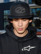 Alpinestars Contender Trucker Hat, sort lifestyle-kasket med poly mesh-bagside, struktureret krone med et broderet Astars logo-mærke, bæres af en mand med en sort Alpinestars hættetrøje, afslappet motorsport-inspireret streetwear.