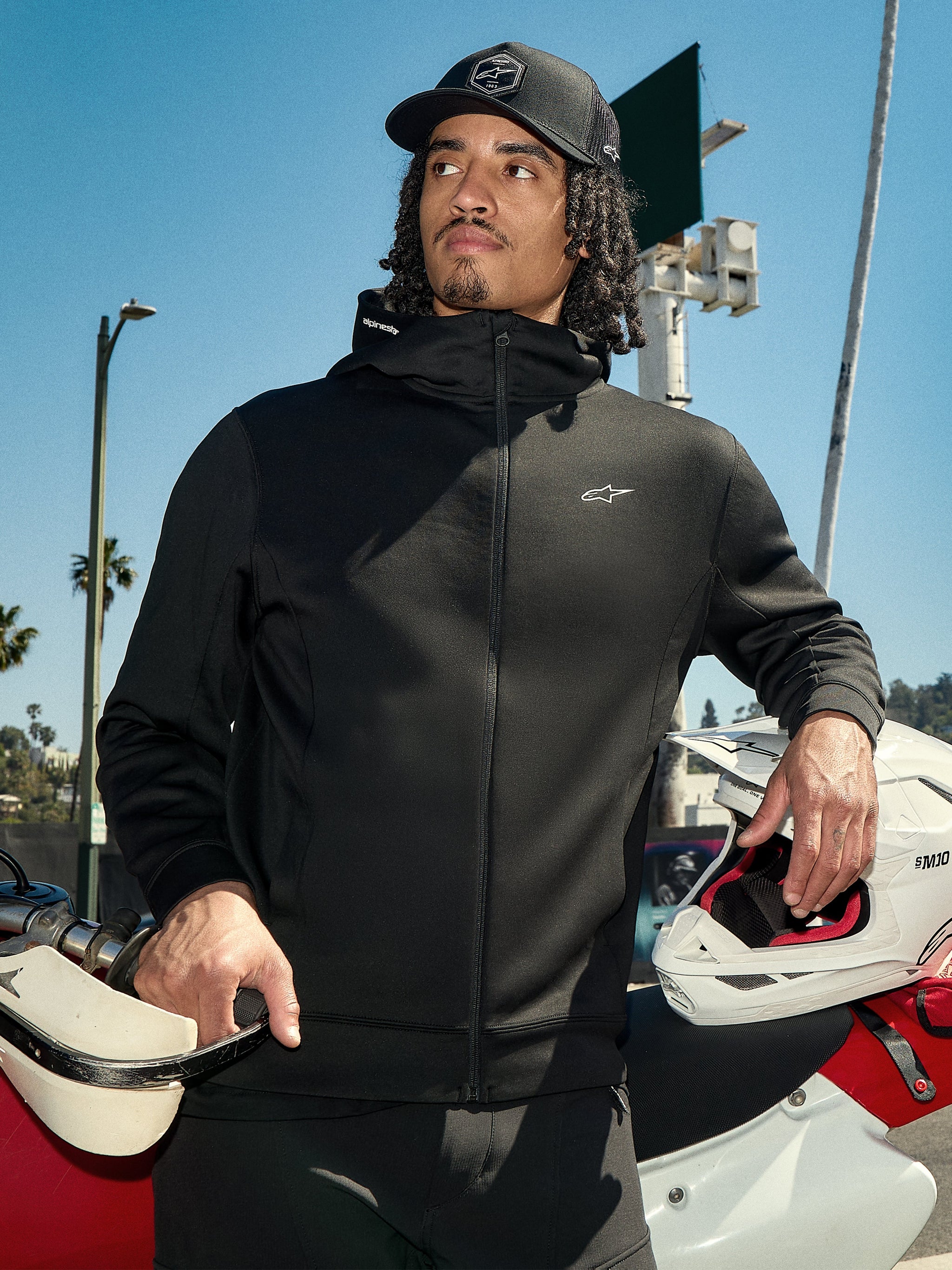 Alpinestars Cypher hættetrøje, sort lifestyle-fleece med lynlås, høj hætte med branding, lille hvidt Astars-logo på brystet, båret med en sort kasket af en kører, der holder en hvid motocrosshjelm, afslappet motorsport-streetwear